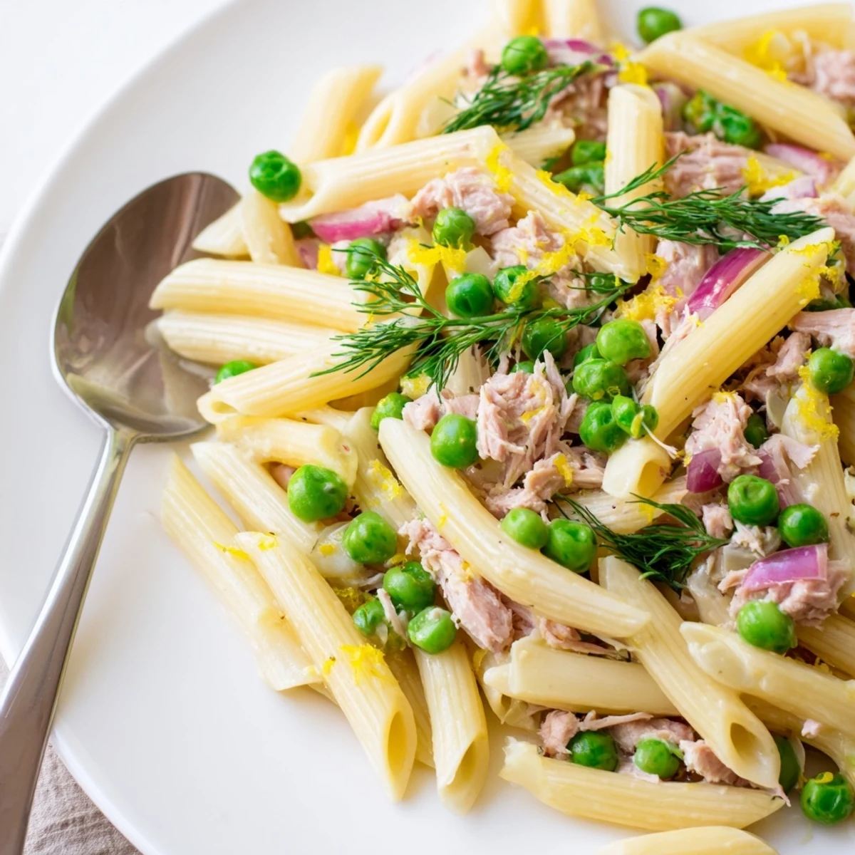 Healthy Lemony Tuna Pasta Salad in a glass serving dish, tossed with parsley, capers, and peas, alongside a chilled glass of white wine on a sunny table.
