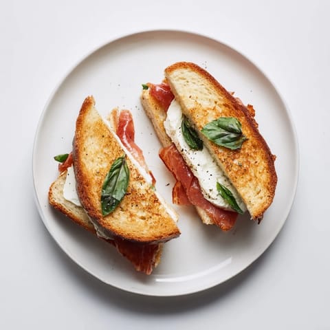 A golden-brown Crispy Prosciutto & Burrata Grilled Cheese on sourdough, served with fresh basil leaves.
