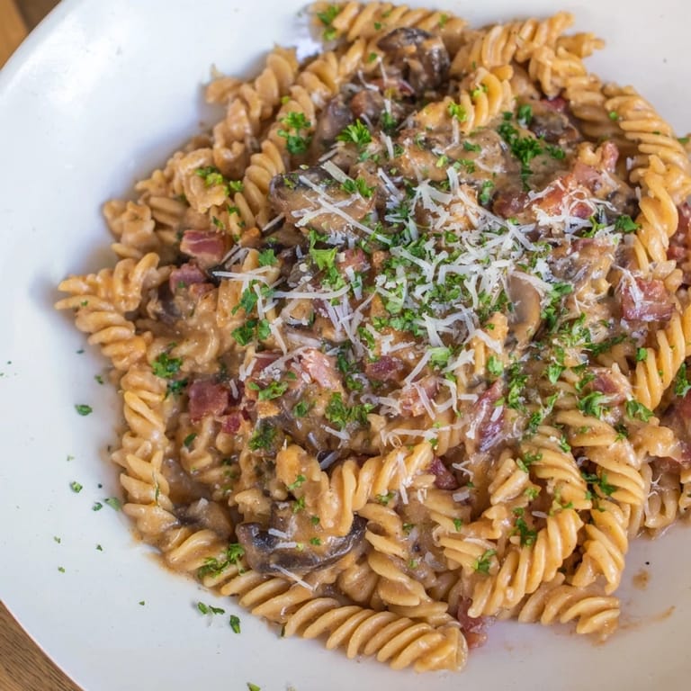 Close-up of golden-brown fusilli with bacon, onions, and mushrooms, plated with Parmesan.